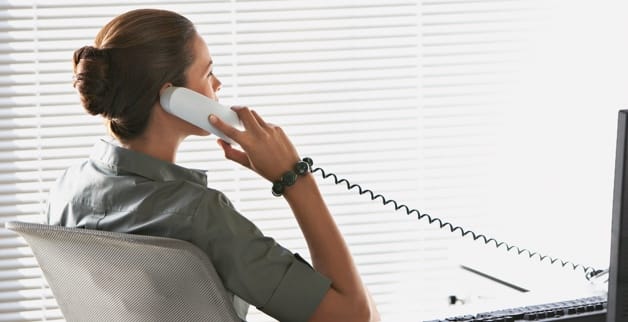 A woman leans back in a chair while speaking on a desk phone. 